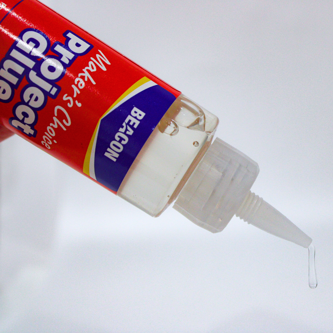 A close-up of Beacon's Project Glue in the bottle with a red and blue label, dispensing a single drop of transparent glue against a white background.